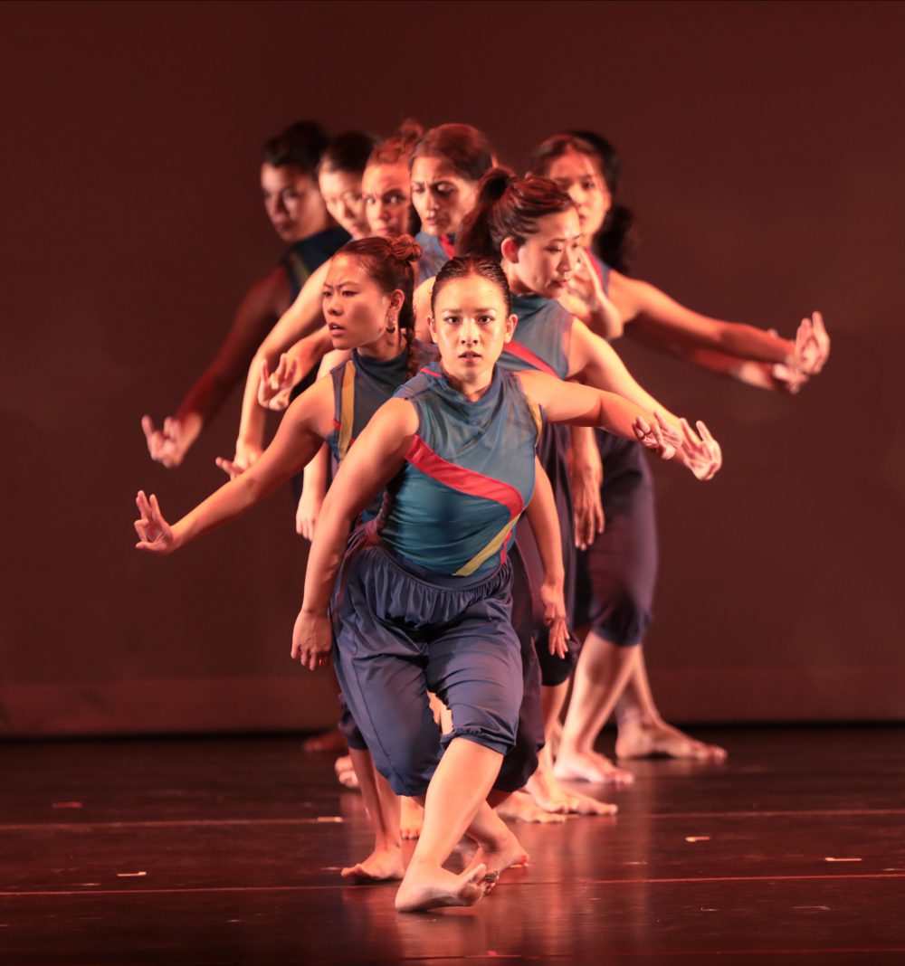 A group of about ten medium-light skinned dancers are in a line on stage. They are all wearing matching outfits that are blue sleeveless shirts with a red and green stripe across, darker blue capri pants. Every other one of them staggered out to a side, with their heads facing down and their palms up. The first person in line is the focus and is staring directly at the camera.