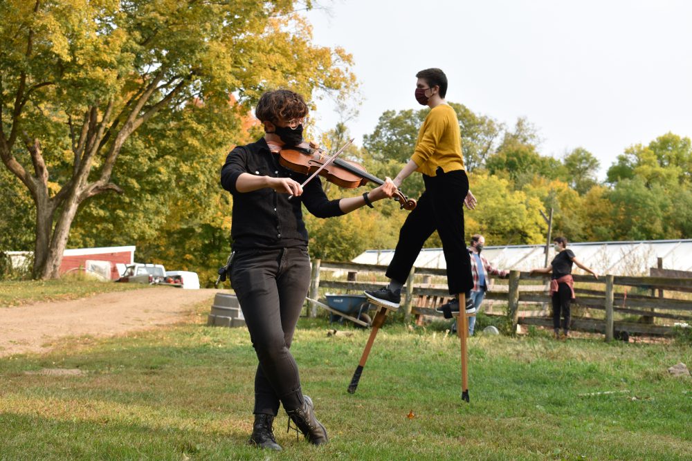 There are two light-skinned people, with short brown hair and wearing medical masks, performing on a barnyard surrounded by yellow and green trees. One of them plays violin while the other walks on stilts behind them.