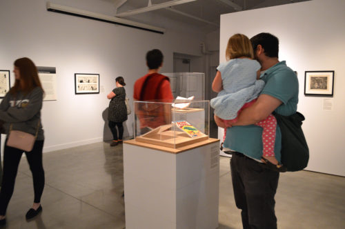In a gallery space with white walls, people look at works of art hung on walls and in cases, in the foreground a man holds a young girl and offers her a closer look
