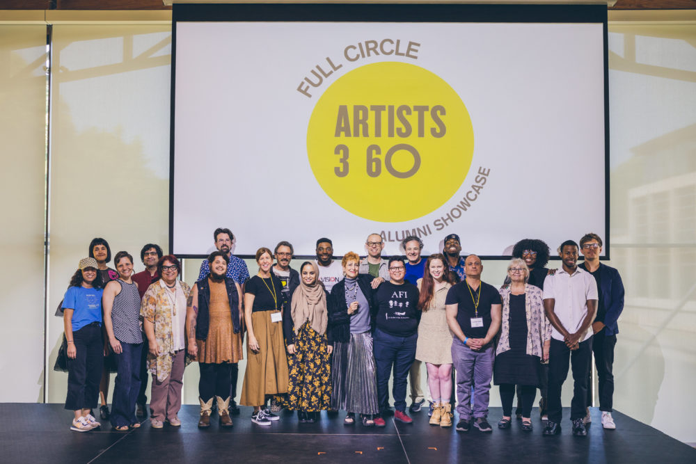 In front of a large screen that says Artists 360 Full Circle stand twenty artists in two rows
