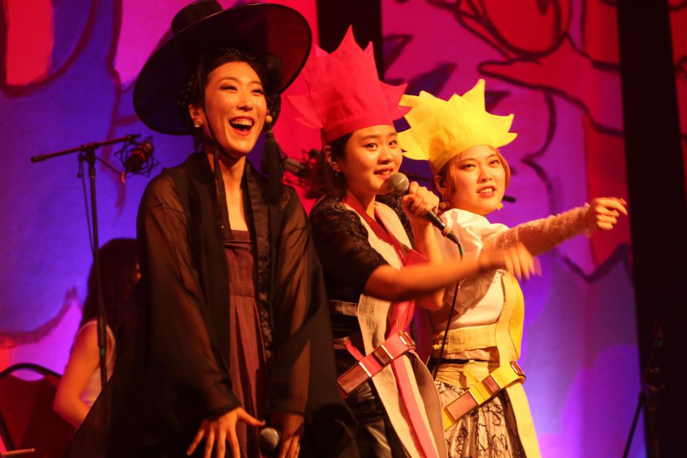Three Korean women in vibrant, updated traditional dress and hats, perform on stage in from of a bright purple and pink background. The women stand in a line close-up, and the artist in the center holds a microphone.