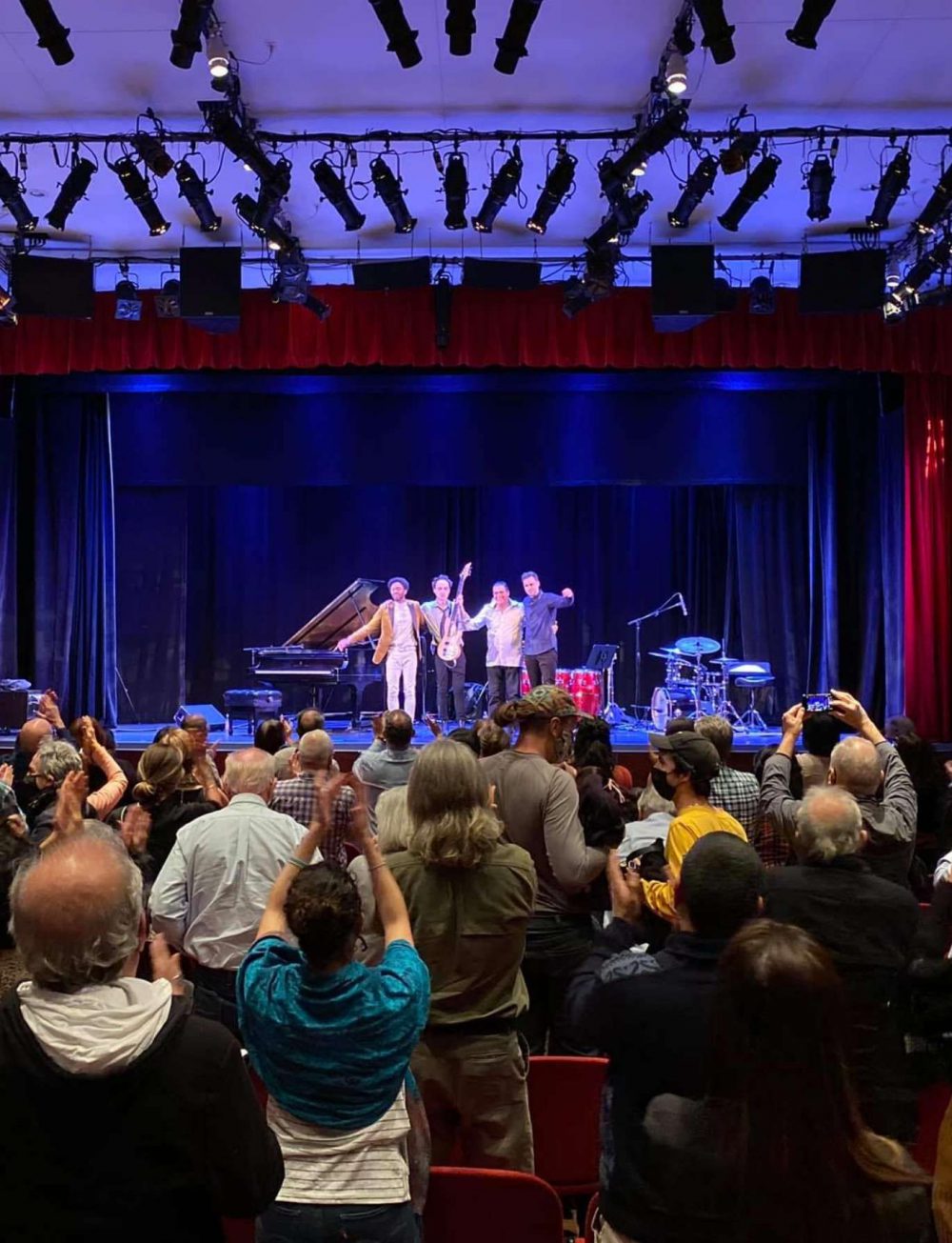 Shot from the back of the performance space, a group of four musicians stand on the stage in a row taking their final bow. They are surrounded by their instruments on a deep blue lit stage and framed with red curtains. The audience stands and claps.