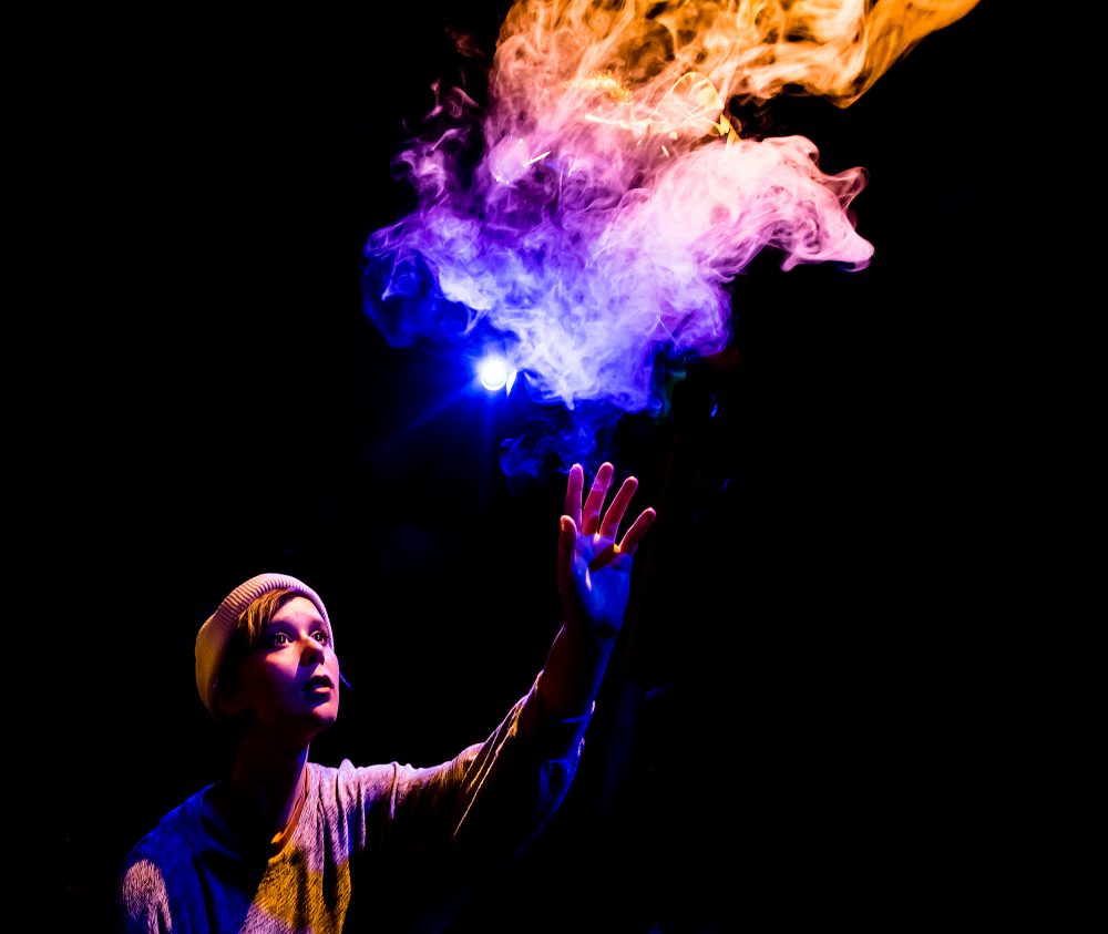 An actor on a darkened stage reaches toward a rainbow swirl of smoke. They were a light-colored long sleeve top and knit cap.