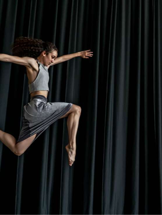 A dancer leaps into the air in front of a grey curtain.