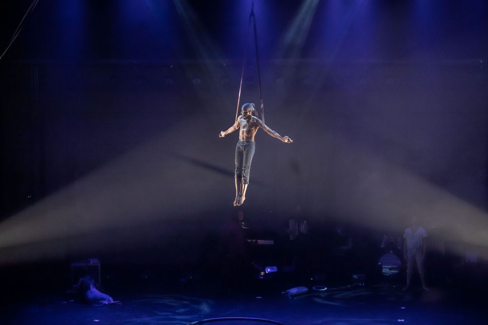 Man suspended in the air in a harness with his arms outstretched. Two lights shine on his body.