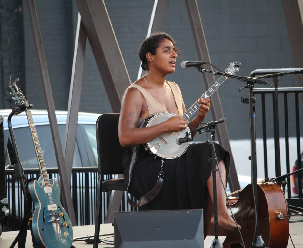 Woman sits on a stage outdoors playing the banjo and singing.