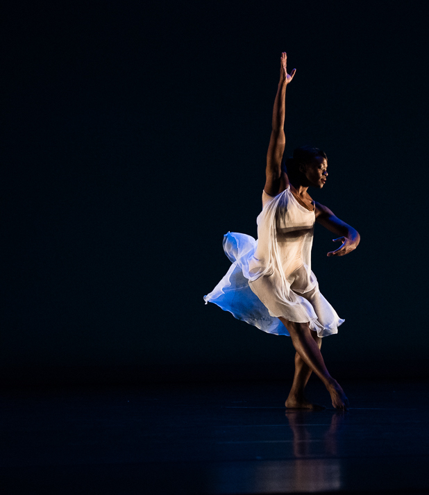 Young black woman wearing an ivory nightgown-like outfit pointing her toes on the ground while raising one arm and holding the other in first position on an illuminated stage