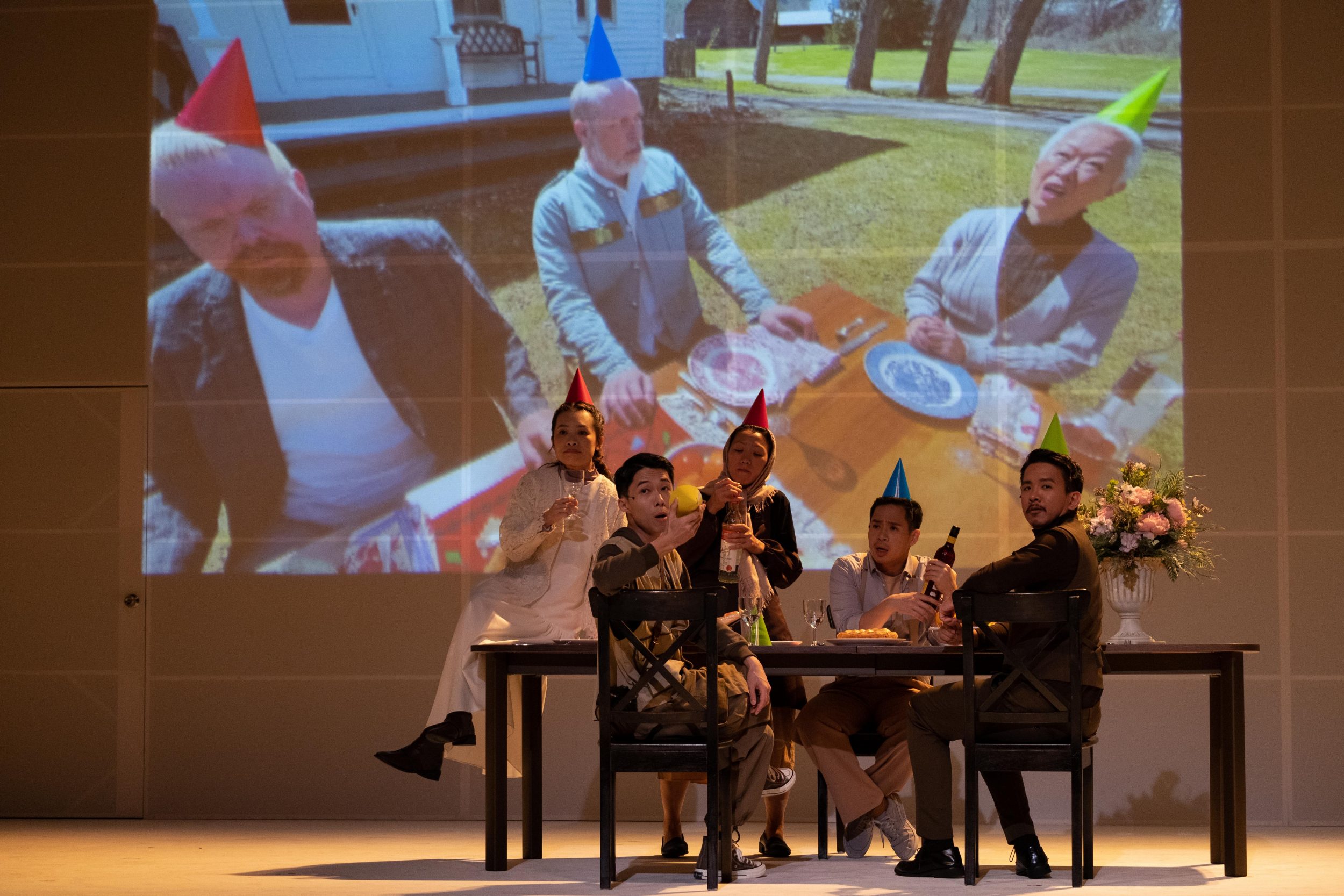 A group of five actors sit at a dining table onstage. Behind them, on a very large screen, a group of three people can be seen. Both groups are hosting a party and all of the cast members wear party hats.